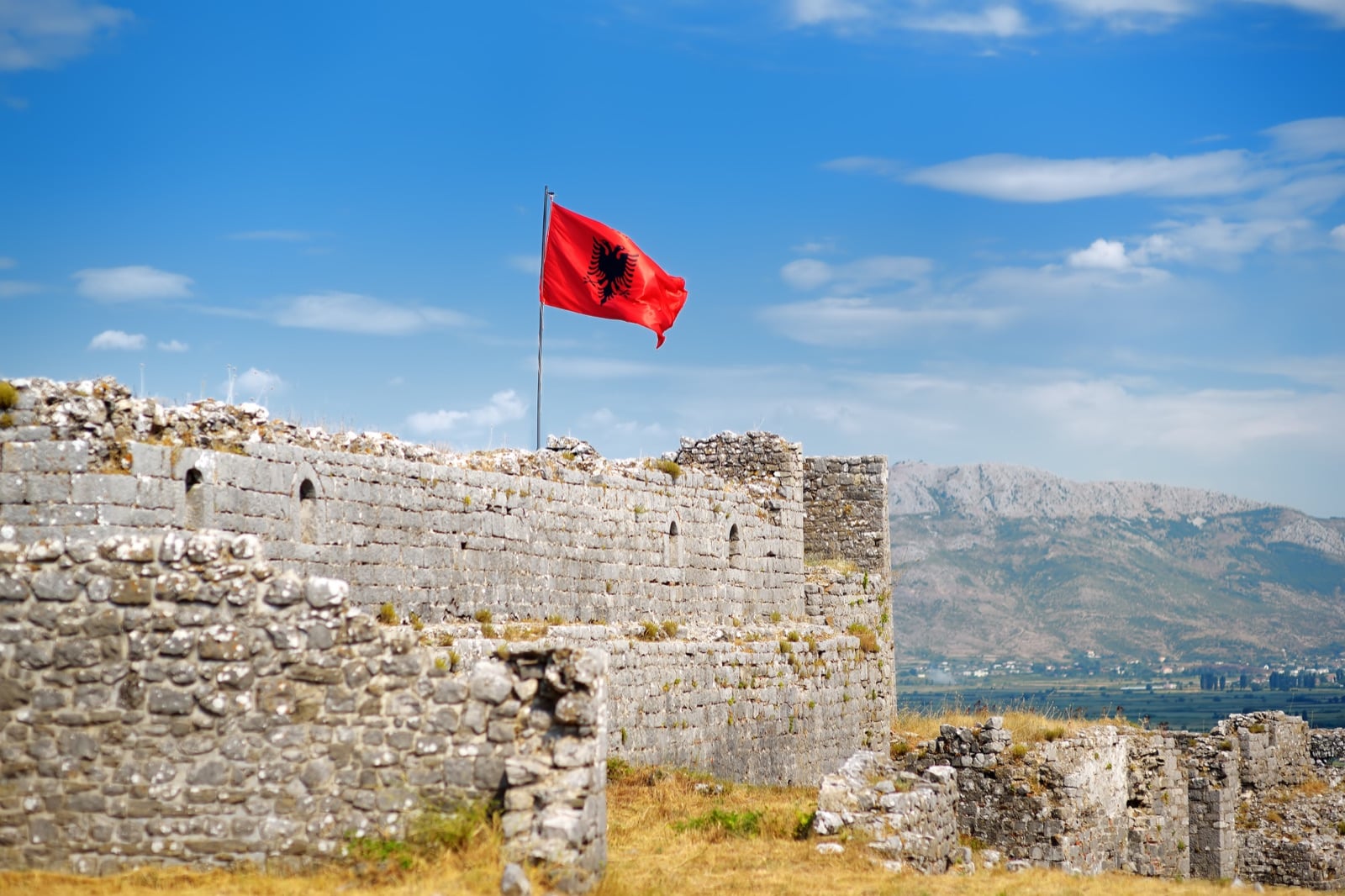 Albanian flag flying over the stone walls of an ancient hilltop fortress with mountains behind